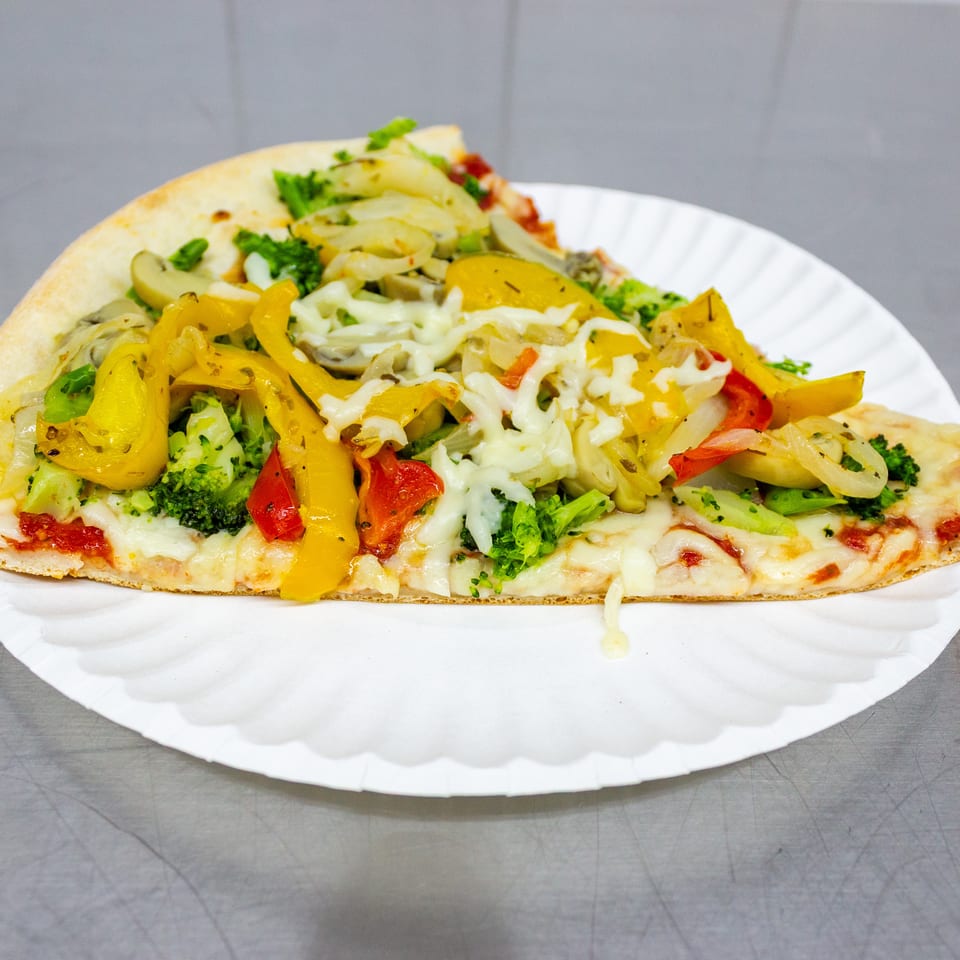 Vegetable pizza topped with melted cheese, broccoli, peppers, mushrooms, onions, and tomatoes on a white paper plate.