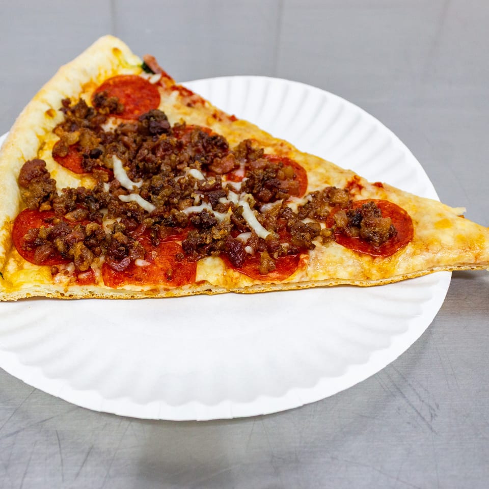 Large triangular pizza slice topped with pepperoni, crumbled meat, melted cheese, and bacon on a white paper plate.