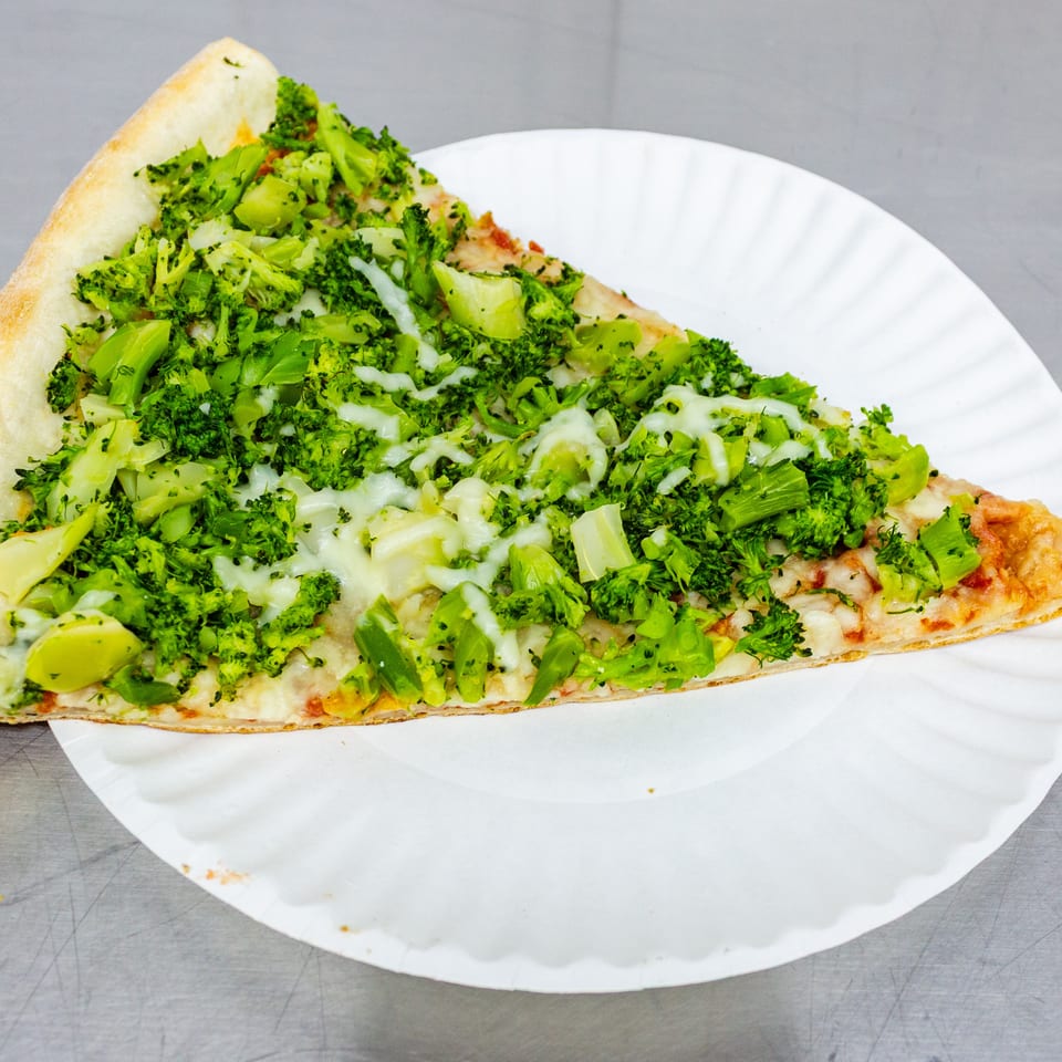 Cheesy pizza slice topped generously with chopped broccoli, served on a white paper plate.