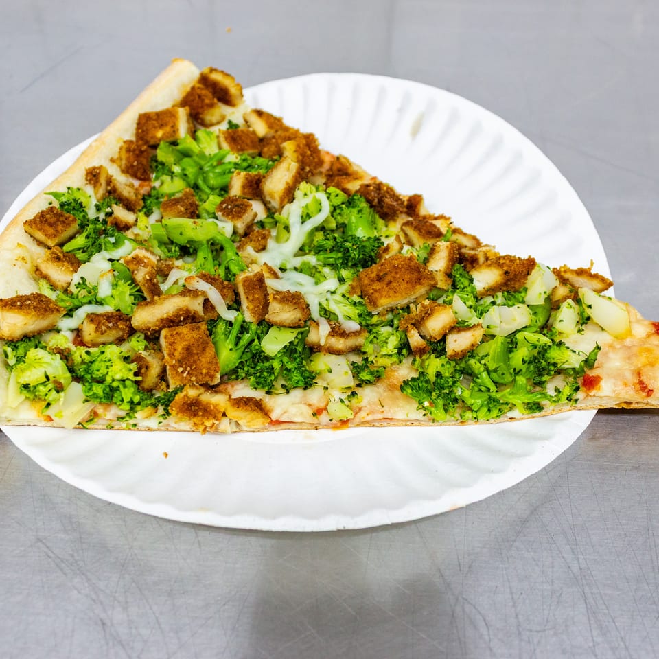 Large rectangular pizza topped with broccoli, breaded chicken pieces, melted cheese, and white sauce on a paper plate.