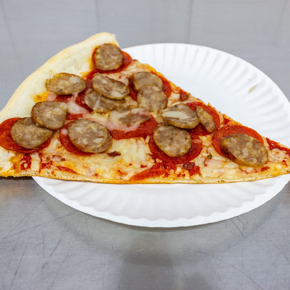 Large triangular pizza slice topped with pepperoni, sliced sausage, melted cheese, and tomato sauce on a white paper plate.